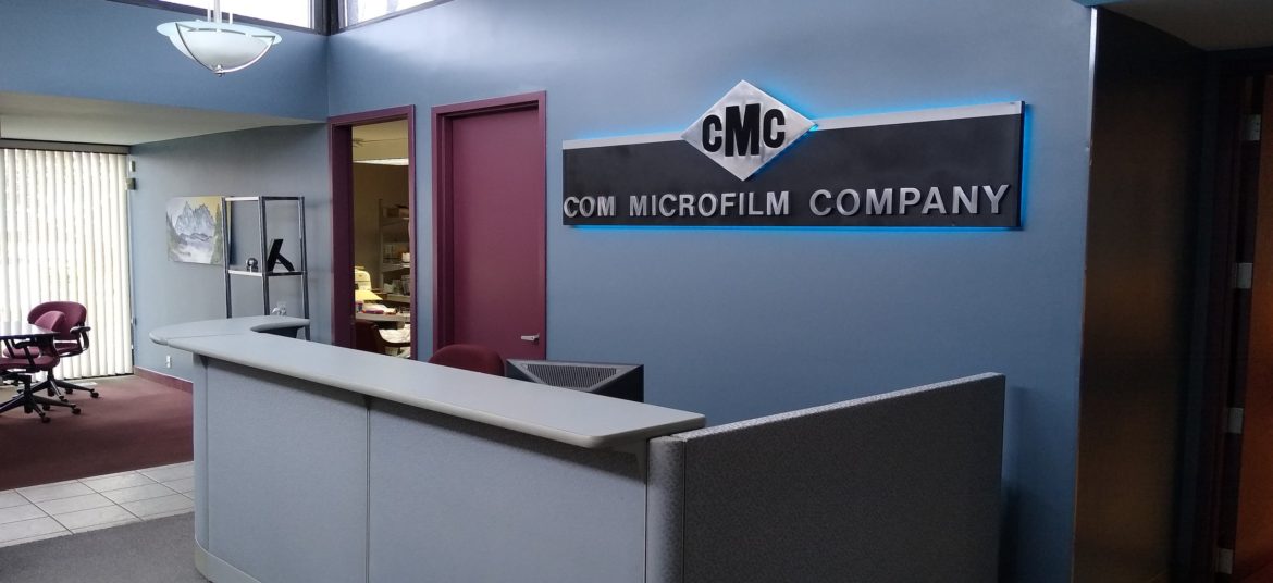 Lobby entrance of CMC Com Microfilm Company with a reception desk, back lighted sign, sky lights and office doors, Board Table in the far left background.